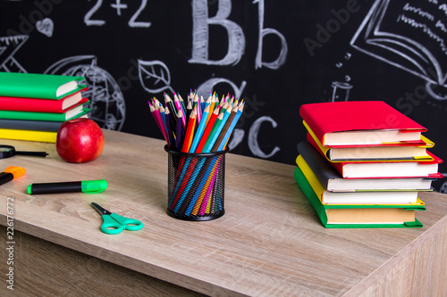 Back to school supplies. Two piles of books, felt-tip pens, magnifying glass, scissors, red apple and a chalkboard on the background