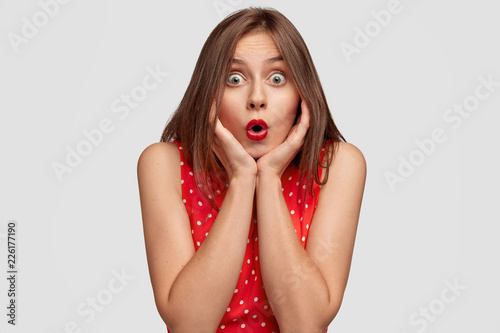 Terrific Caucasian woman opens mouth widely, holds breath, keeps mouth rounded, holds chin with both hands, wears red lipstick, cant believe in shocking news, isolated over white background.