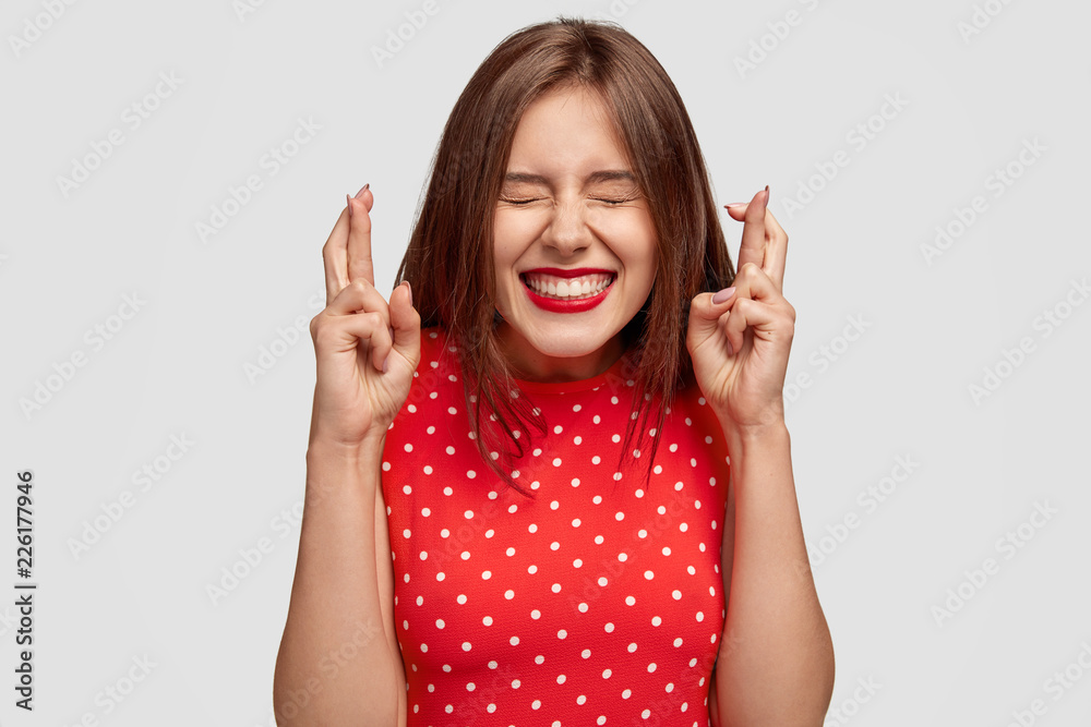 Pleased attractive European woman makes wish to win, raises hands with crossed fingers, waits for lottery results, closes eyes, has red lips, dressed in fashionable dress, isolated over white wall Pleased attractive European woman makes wish to win, raises hands with crossed fingers, waits for lottery results, closes eyes, has red lips, dressed in fashionable dress, isolated over white wall