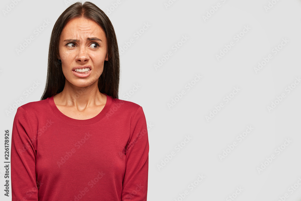 Beautiful displeased woman frowns face in displeasure and with aversion, doesnt like something, dressed in casual red sweater, poses over white blank wall with free space for your promotion. Beautiful displeased woman frowns face in displeasure and with aversion, doesnt like something, dressed in casual red sweater, poses over white blank wall with free space for your promotion.