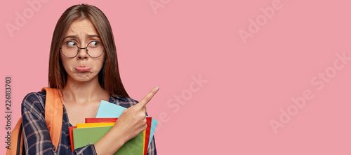 Studio shot of dissatisfied European woman has dejected facial expression, indicates with index finger at upper right corner, carries rucksack, textbooks, feels offended, isolated over pink background