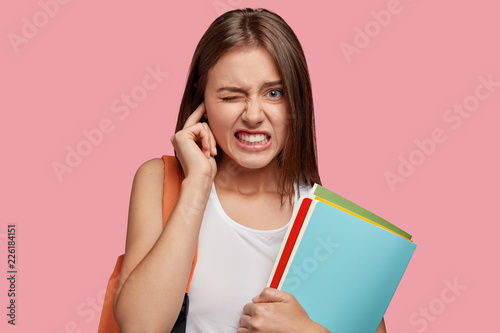 Displeased European girlfriend being annoyed with unpleasant sound, plugs ear, clenches teeth with negativity, feels irritated, hears noise, poses against pink background, holds folders in hands