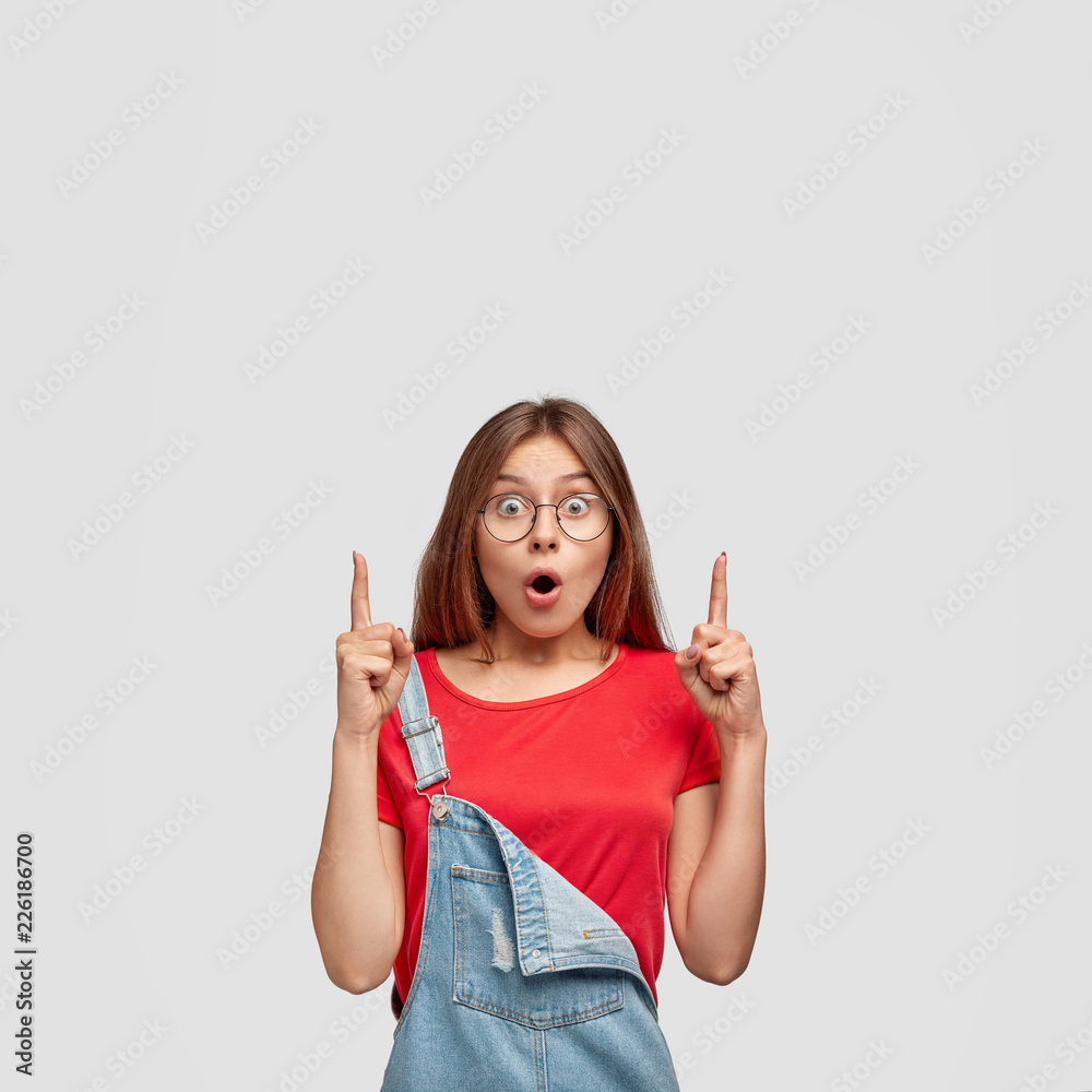 Astonished cute European teenage girl with dark straight hair, healthy skin, dressed in casual clothes, points with both index fingers upwards, shows something unbelievable, models in studio Astonished cute European teenage girl with dark straight hair, healthy skin, dressed in casual clothes, points with both index fingers upwards, shows something unbelievable, models in studio