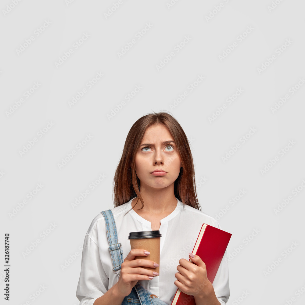 Indoor shot of displeased gloomy student holds disposable cup of coffee, notepad, looks with bewilderment upwards, frowns eyebrows, free space above for your promotional content. Learning concept Indoor shot of displeased gloomy student holds disposable cup of coffee, notepad, looks with bewilderment upwards, frowns eyebrows, free space above for your promotional content. Learning concept