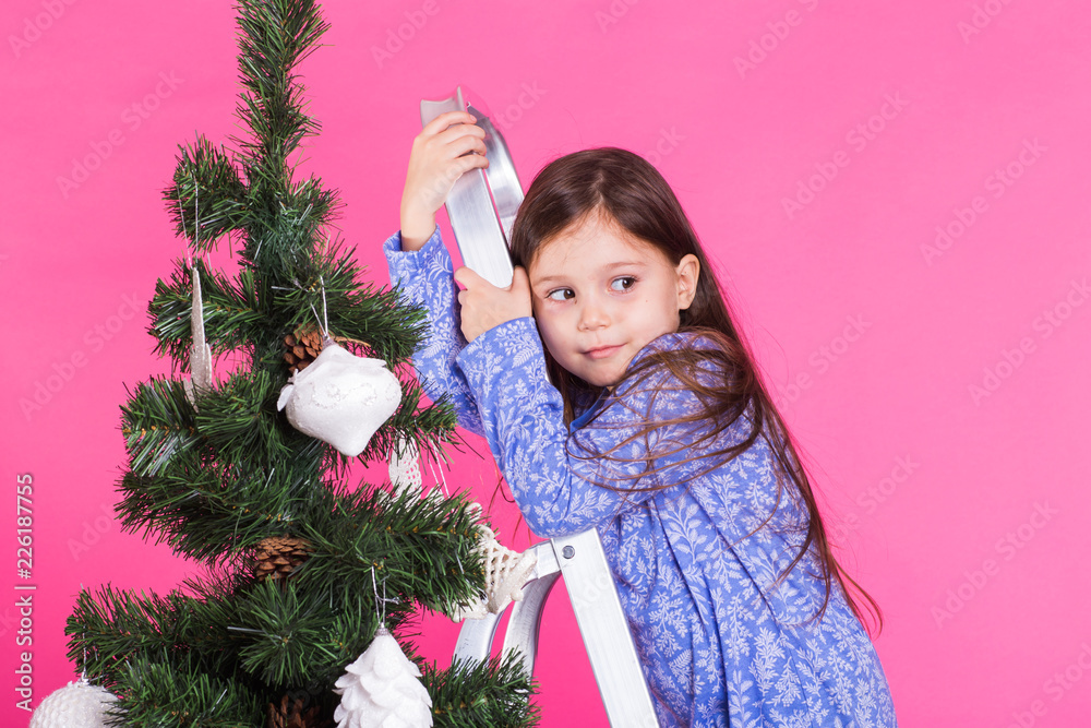 Children, holidays and christmas concept - little girl near christmas tree on pink background Children, holidays and christmas concept - little girl near christmas tree on pink background
