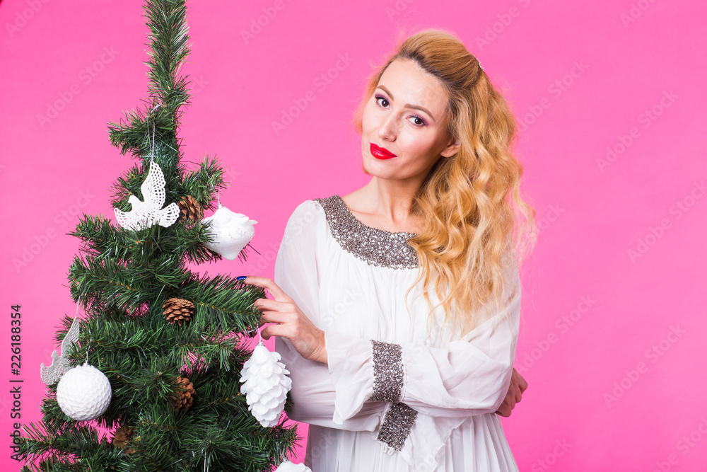 People, holidays and christmas concept - young blonde woman standing near christmas tree on pink background