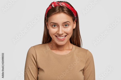 Photo of pleased young female model with toothy expression, looks joyfully in...