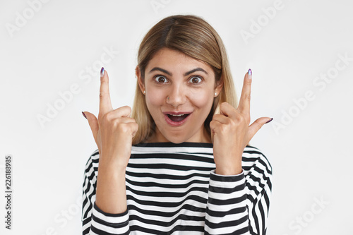 Look at this. Pretty female wearing striped top having ecstatic overjoyed look, amazed with something, indicating copyspace on blank studio wall upwards, pointing raised index fingers and smiling