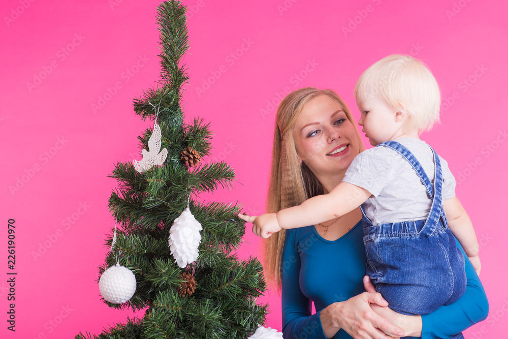Holidays, family and christmas concept - young mom with her little daughter near christmas tree on pink background Holidays, family and christmas concept - young mom with her little daughter near christmas tree on pink background