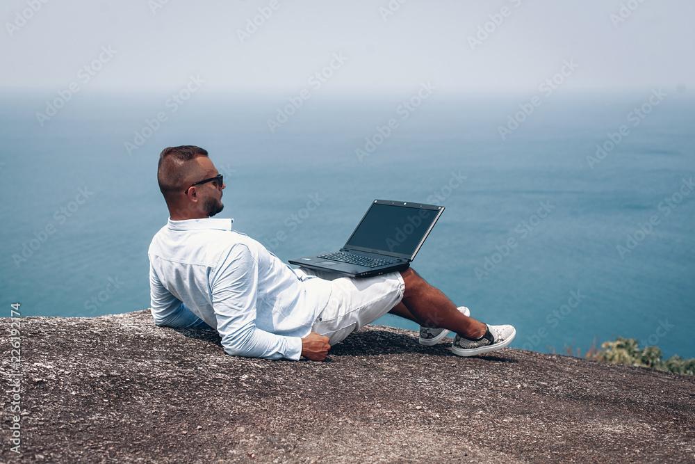 young guy work on laptop on breach .  beautiful sea view. young businessman in sunglasses blue shirt and shorts working with laptop on the rocks