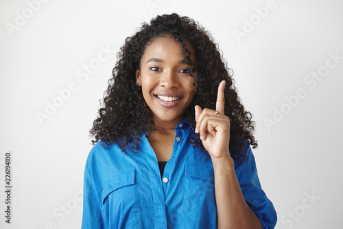 I have an idea. Cheerful cute African student girl with voluminous hair smiling excitedly and raising fore finger, full of great ideas while working on her home assignment, thinking outside the box