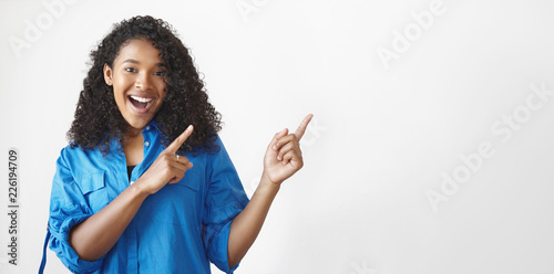 Look at this. Horizontal shot of emotional gorgeous joyful young African American female having excited fascinated facial expression, keeping mouth wide opened and pointing fore fingers upwards photo