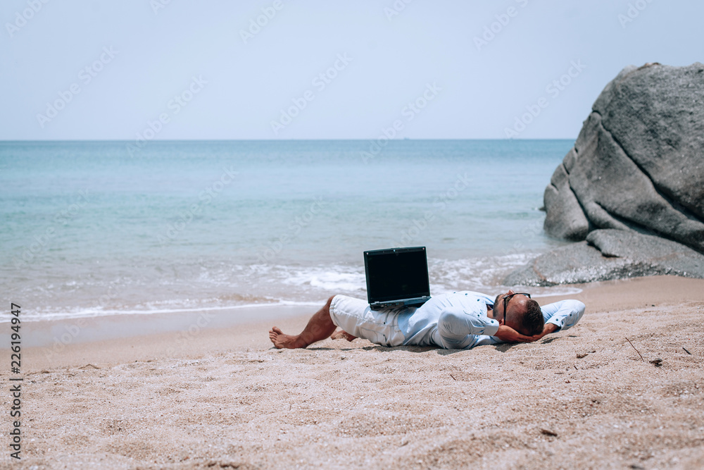 young businessman in sunglasses blue shirt and shorts working with laptop on the beach