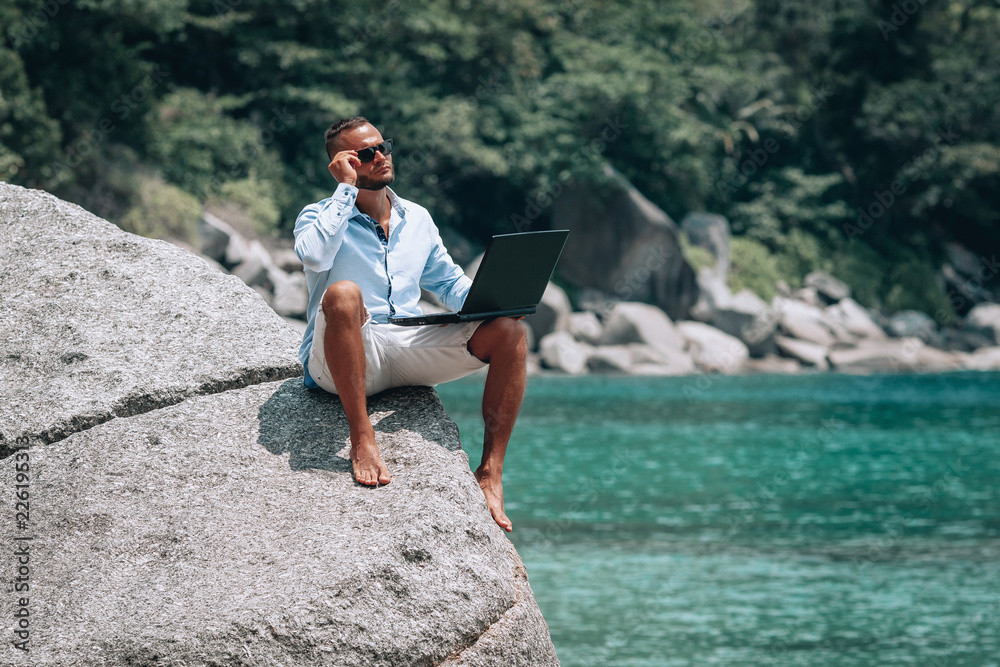 young businessman in sunglasses blue shirt and shorts working with laptop on the beach