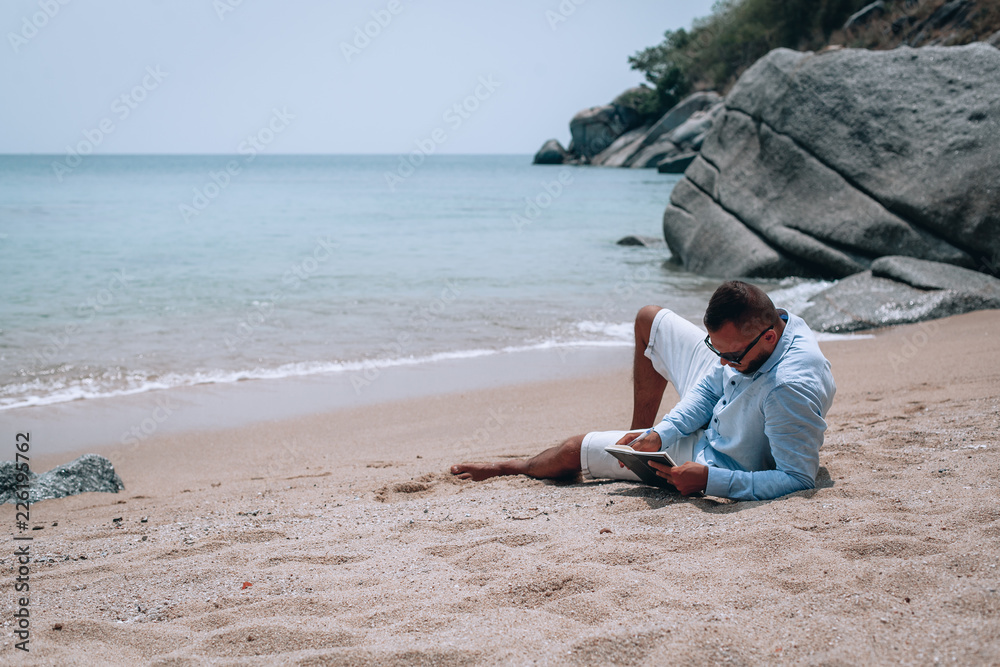 a young businessman in sunglasses, blue shirt and shorts writes in a notebook