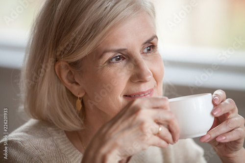 Thoughtful happy middle aged senior woman with beautiful face looking away drinking morning coffee  smiling mature old lady holding tea cup relaxing with positive thoughts dreaming enjoying wellbeing
