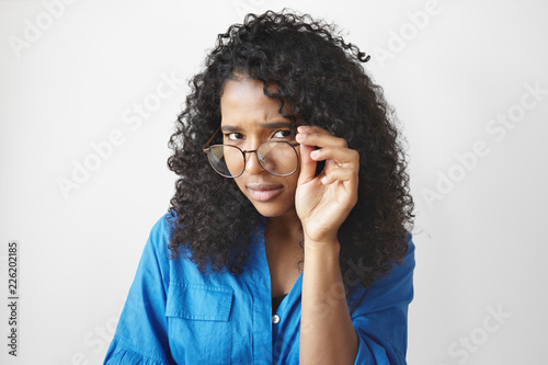 Close up studio shot of beautiful stylish young African American woman in blue shirt holding trendy eyewear and looking at you with scrutinizing facial expression, having strict suspicious look