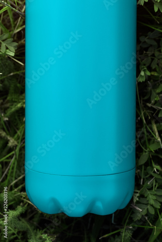 Stainless thermos water bottle, light blue color. Isolated on green grass background with sunlight effect.