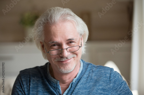Charming middle aged elderly old grey haired man wearing glasses looking at camera posing at home, happy handsome positive single mature senior retired male with smiling face head shot portrait