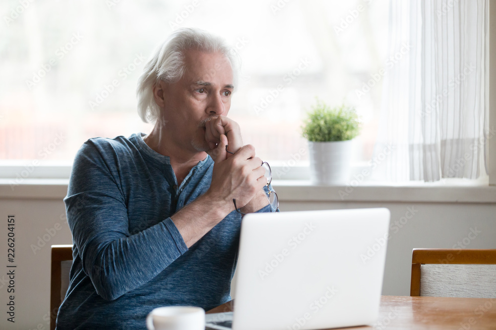 Serious thoughtful mature senior man looking away thinking of online problem solution near laptop, focused worried old middle aged male concerned feeling anxious or perplexed sitting with computer Serious thoughtful mature senior man looking away thinking of online problem solution near laptop, focused worried old middle aged male concerned feeling anxious or perplexed sitting with computer