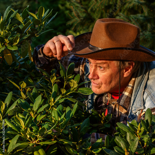 Senior adult man or a farmer, in a brown cowboy hat, looks out of the bushes and makes a grimace. Concept of active leisure for middle-aged and older people. For instagram format. Square.