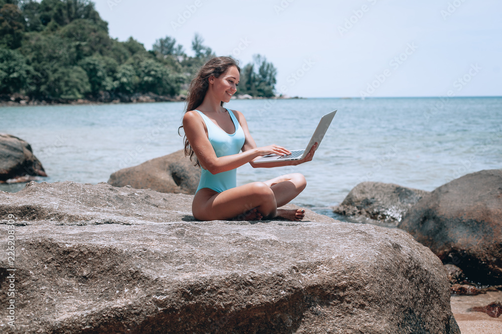 Young cute woman using laptop and sitting on stone near sea, empty morning beach. Concept of modern technology and working on nature, summer vacations.