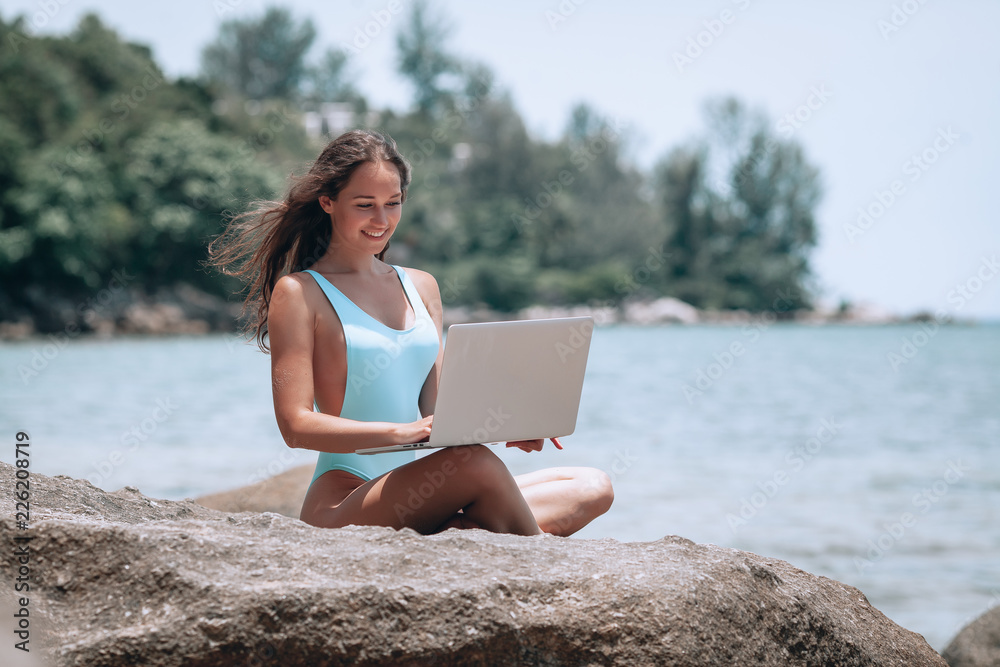 Young beatiful woman working with laptop on empty beach and sitting on stone, showing thumbs up. Concept of resting on morning sea and summer vacations, modern technology. Young beatiful woman working with laptop on empty beach and sitting on stone, showing thumbs up. Concept of resting on morning sea and summer vacations, modern technology.