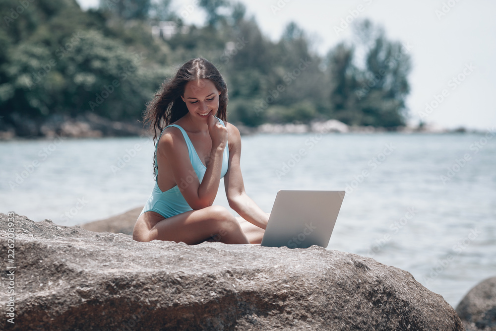 Young Caucasian woman working with laptop on beautiful beach and sitting on white sand . The concept of rest on the morning sea and summer holidays, modern technology.