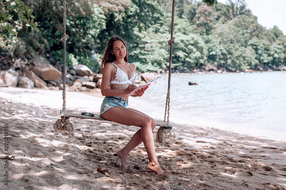 Young fit girl in denim shorts and a white bra swinging on a swing on the  shore of the Andaman sea in Phuket. that the writes and reads in Notepad  Stock Photo