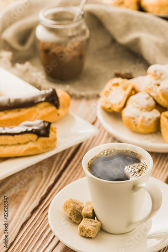 Home cooked profiteroles and eclairs with a cup of black coffee on a wooden table.