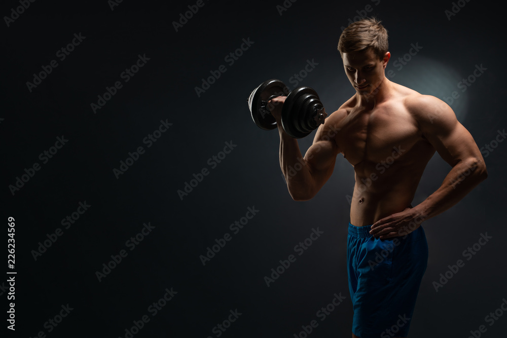Handsome power athletic man in training swap muscles with dumbbells. A strong six-pack bodybuilder, perfect abs, shoulders, biceps, triceps and chest. On a dark background under the banner