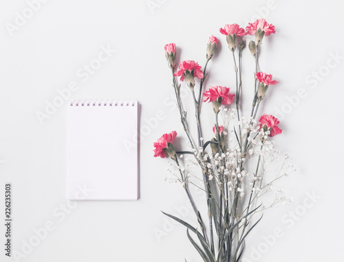 Pink cloves flowers and gypsophila next to small white notebook isolated on light gray background. Copy space. flat lay  top view