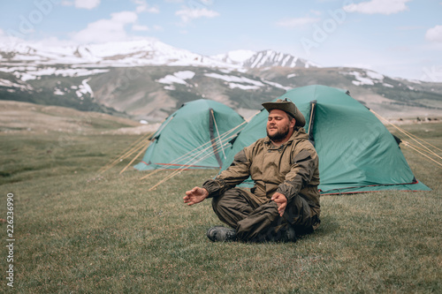 Man with a beard sitting on the grass and meditating. Male doing yoga on mountains near the tents..