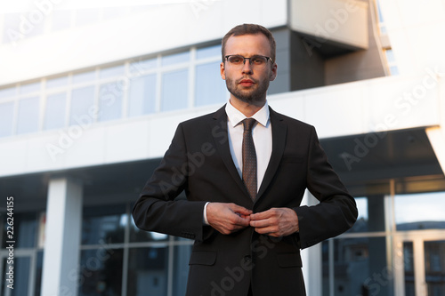 Young businessman in official black suit is standing in front of office building
