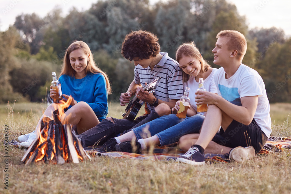 Joyful teenagers sit closely to each other on ground near fire, have picnic together, play acoustic guitar, celebrate something, have outdoor party, drink cold energetic drinks. Leisure concept