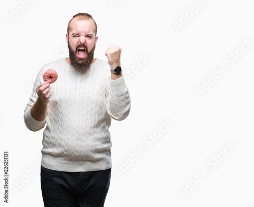 Young caucasian hipster man eating sweet donut over isolated background annoyed and frustrated shouting with anger, crazy and yelling with raised hand, anger concept