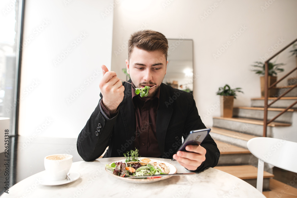 Business man use a smartphone during breakfast in the restaurant. Attractive business man sits in a cozy restaurant, eating salad greens and using a smartphone. Business man use a smartphone during breakfast in the restaurant. Attractive business man sits in a cozy restaurant, eating salad greens and using a smartphone.