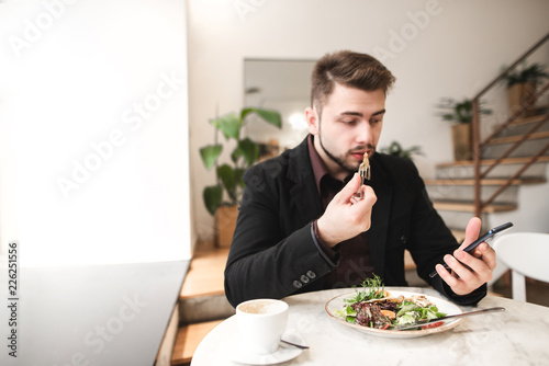 Attractive man in a suit sits in a cozy restaurant  eating salad with a plate and using a smartphone. Business man use a smartphone during lunch in the restaurant. Focus on the smartphone. Copyspace