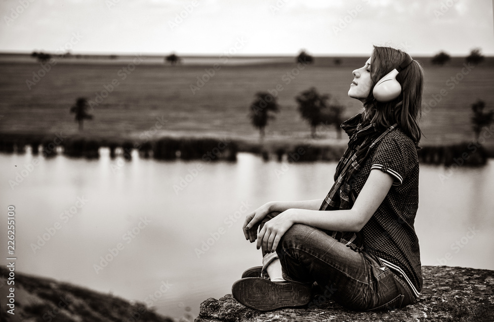 Young fashion girl with headphones sitting near river at countryside. Image in black and white color
