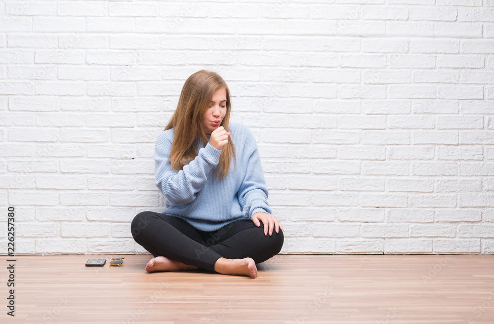 Young adult woman sitting on the floor in autumn over white brick wall feeling unwell and coughing as symptom for cold or bronchitis. Healthcare concept. Young adult woman sitting on the floor in autumn over white brick wall feeling unwell and coughing as symptom for cold or bronchitis. Healthcare concept.