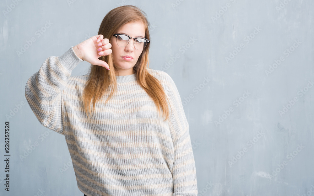 Young adult woman over grey grunge wall wearing glasses with angry face, negative sign showing dislike with thumbs down, rejection concept