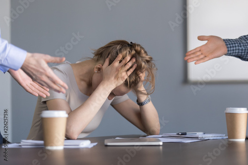 Stressed young businesswoman holding head in hands suffering from unfair gender discrimination at work concept, female employee frustrated by male colleagues bullying humiliating woman at workplace