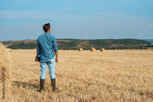 Man farmer agronomist in jeans and shirt stands back in the field after haymaking, with tablet looking into the distance. Rural business, agricultural industry, freedom after work, concept © Руслан Галиуллин