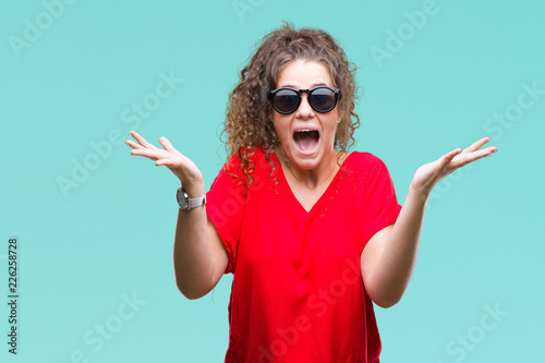 Beautiful brunette curly hair young girl wearing sunglasses over isolated background celebrating crazy and amazed for success with arms raised and open eyes screaming excited. Winner concept