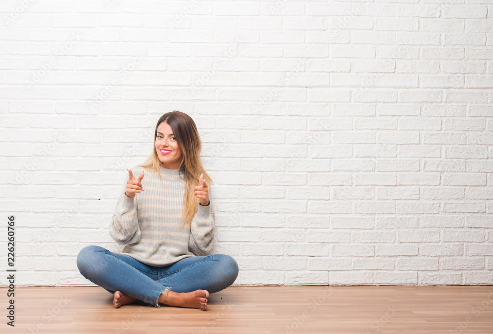 Young adult woman sitting on the floor over white brick wall at home pointing fingers to camera with happy and funny face. Good energy and vibes. Young adult woman sitting on the floor over white brick wall at home pointing fingers to camera with happy and funny face. Good energy and vibes.