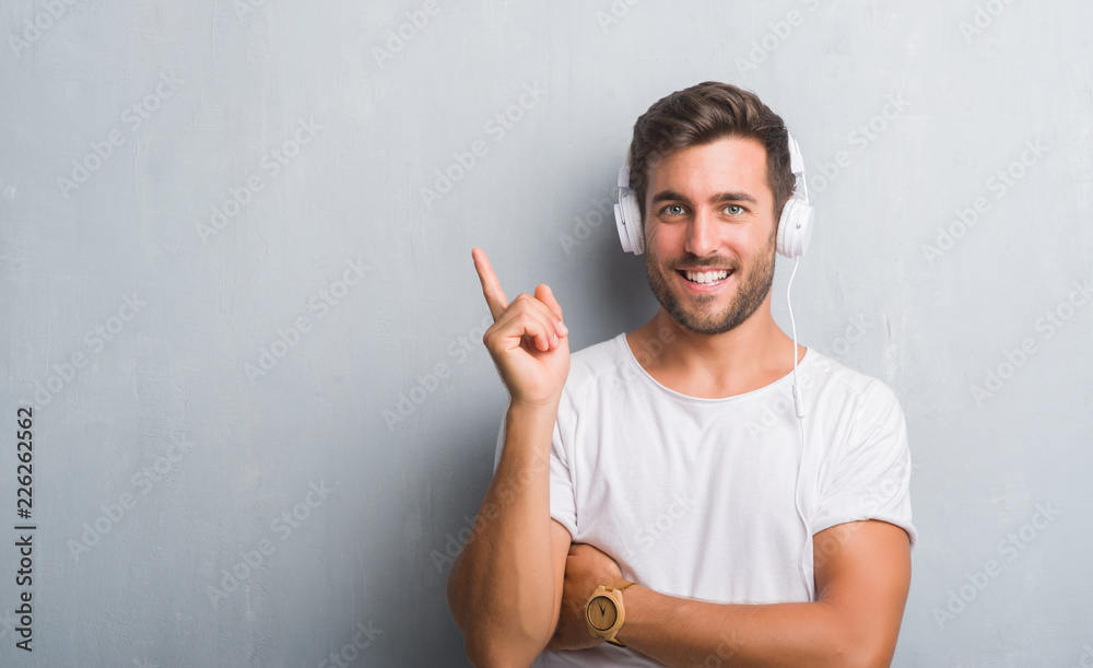 Handsome young man over grey grunge wall wearing headphones listening to music very happy pointing with hand and finger to the side Handsome young man over grey grunge wall wearing headphones listening to music very happy pointing with hand and finger to the side