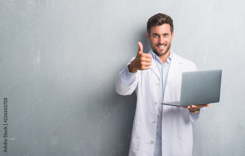 Handsome young man over grey grunge wall wearing white coat using laptop happy with big smile doing ok sign, thumb up with fingers, excellent sign Handsome young man over grey grunge wall wearing white coat using laptop happy with big smile doing ok sign, thumb up with fingers, excellent sign
