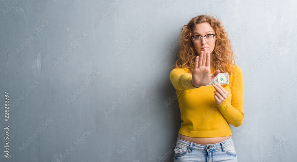 Young redhead woman over grey grunge wall holding a dollar with open hand doing stop sign with serious and confident expression, defense gesture Young redhead woman over grey grunge wall holding a dollar with open hand doing stop sign with serious and confident expression, defense gesture
