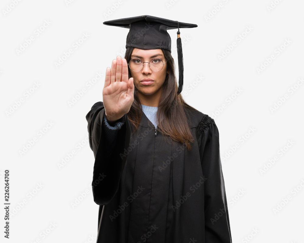 Young hispanic woman wearing graduated cap and uniform doing stop sing with palm of the hand. Warning expression with negative and serious gesture on the face. Young hispanic woman wearing graduated cap and uniform doing stop sing with palm of the hand. Warning expression with negative and serious gesture on the face.