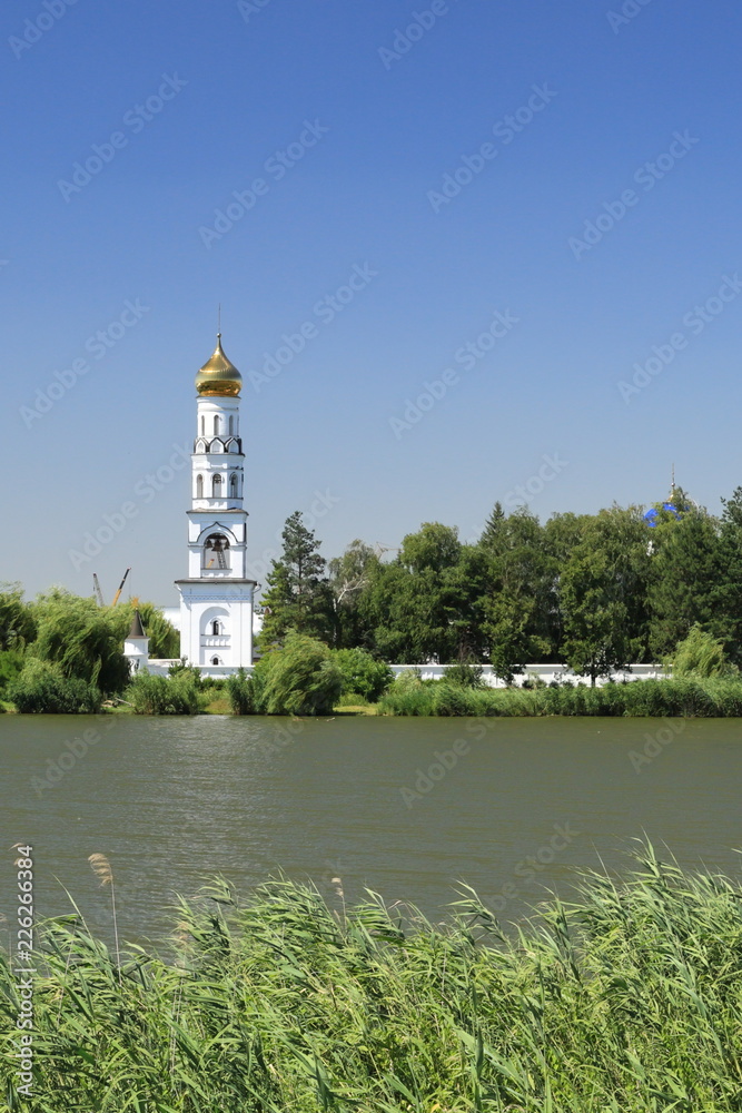 High white bell tower standing on the shore of the lake among the trees